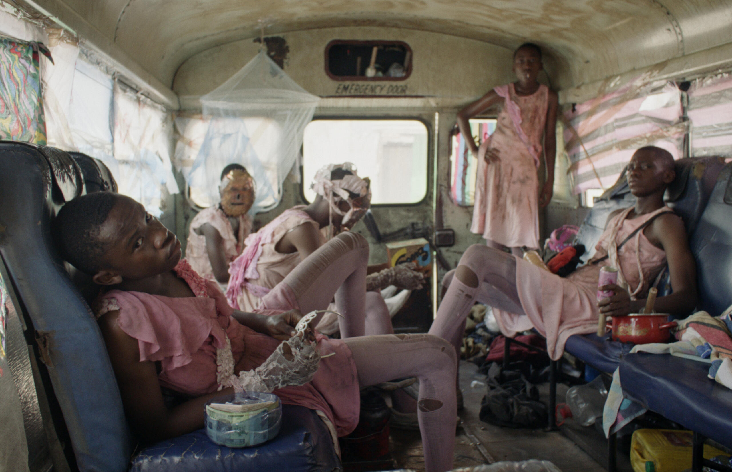 Five people sit or stand in torn and dirty pink clothes in a littered bus. The man in the foreground holds a small crown in his hand.