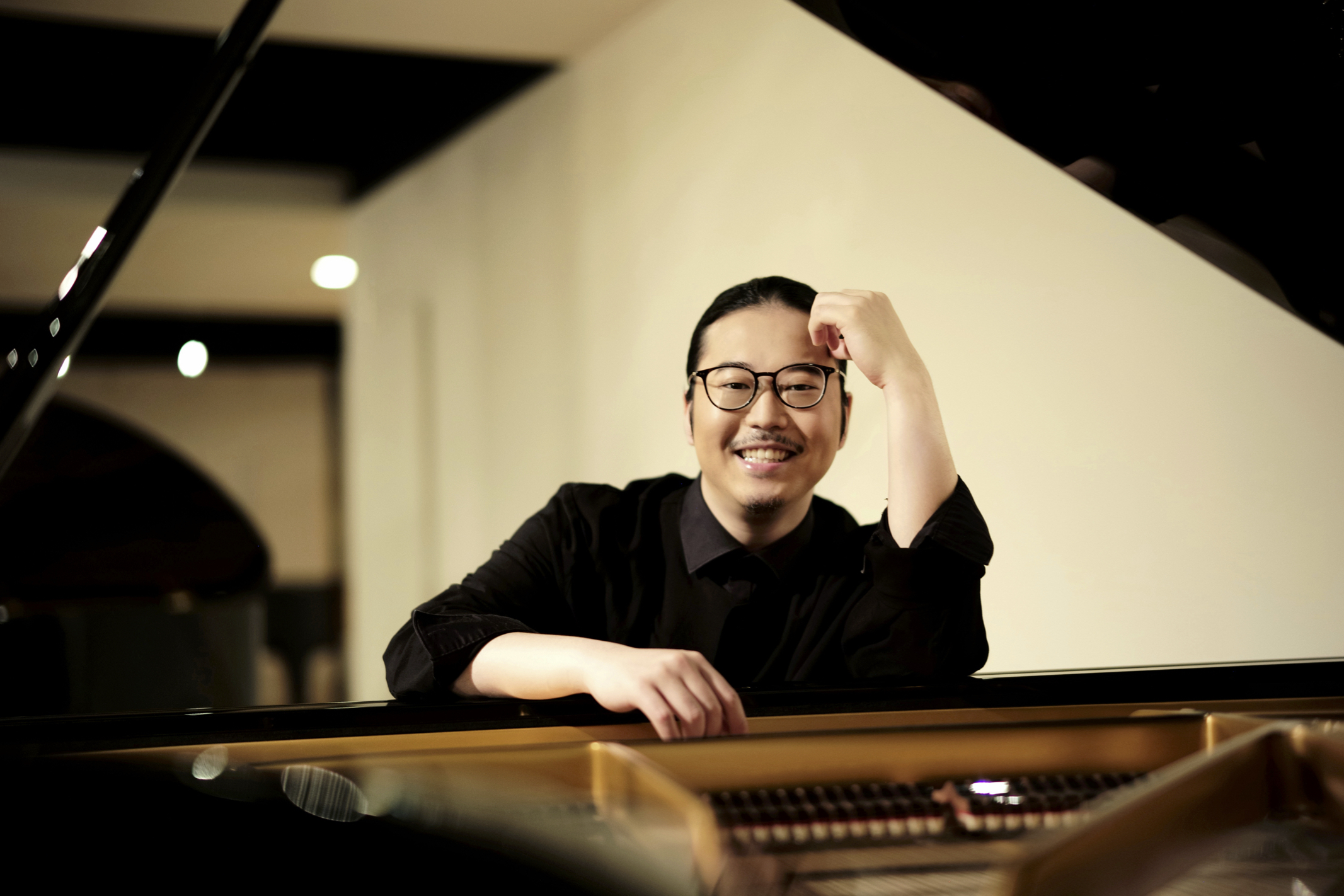 Porträt des Pianisten Kyohei Sorita. Er trägt ein schwarzes Hemd, sitzt hinter einem Flügel und lächelt in die Kamera.