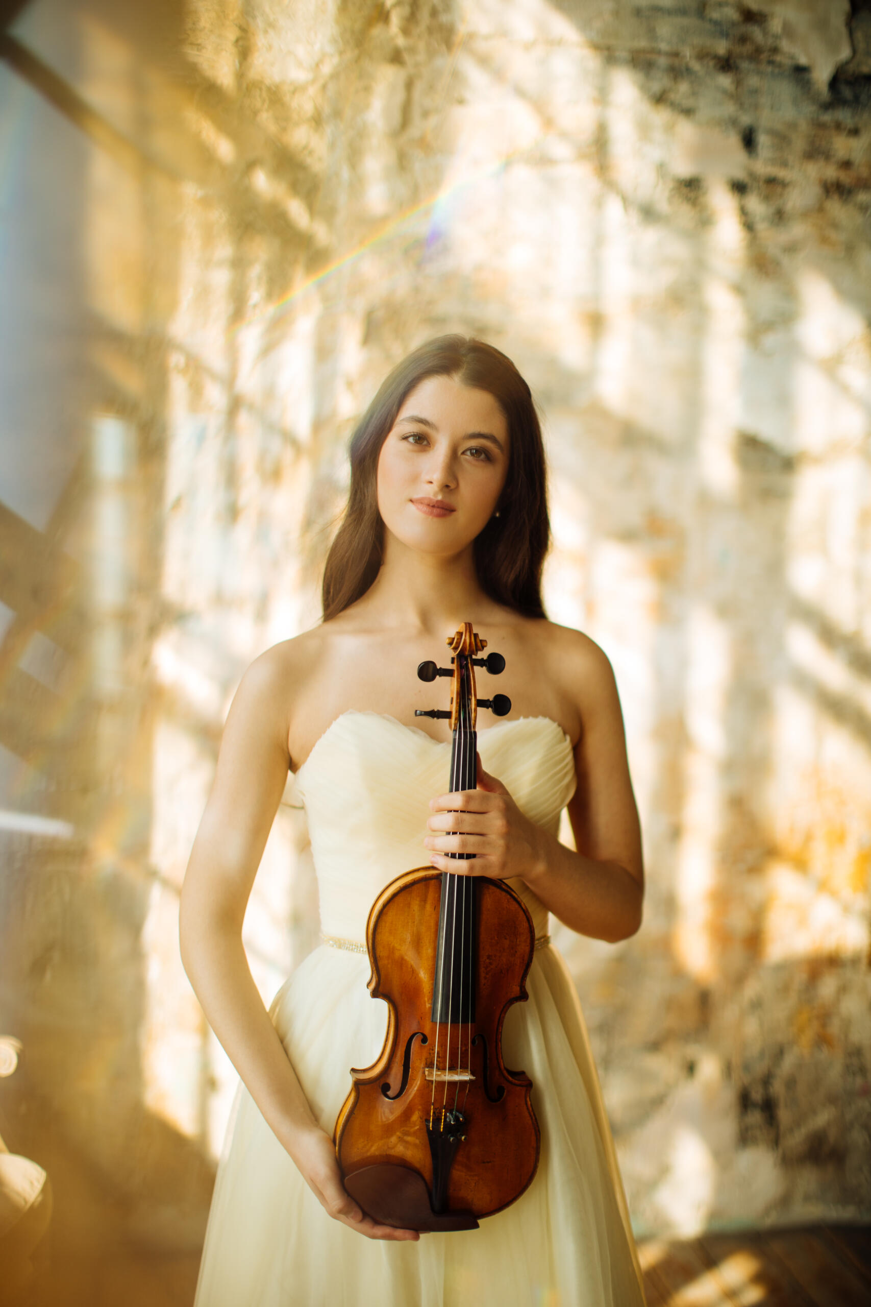 Portrait of the violinist María Dueñas. She wears a beige dress, holds her violin in her hand and stands in a light-flooded room.