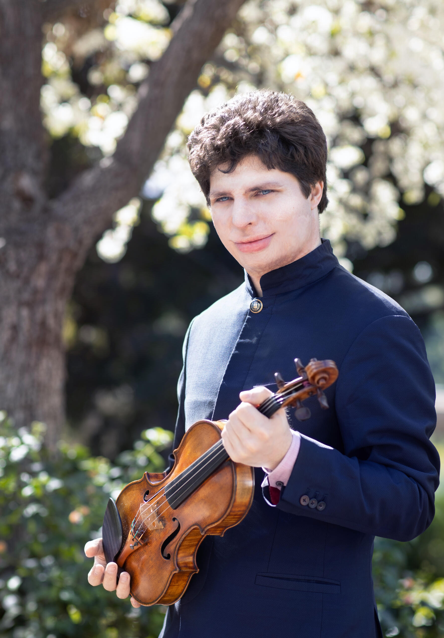Portrait of the violinist Augustin Hadelich. He is wearing a blue suit jacket and is standing in a garden with his violin.