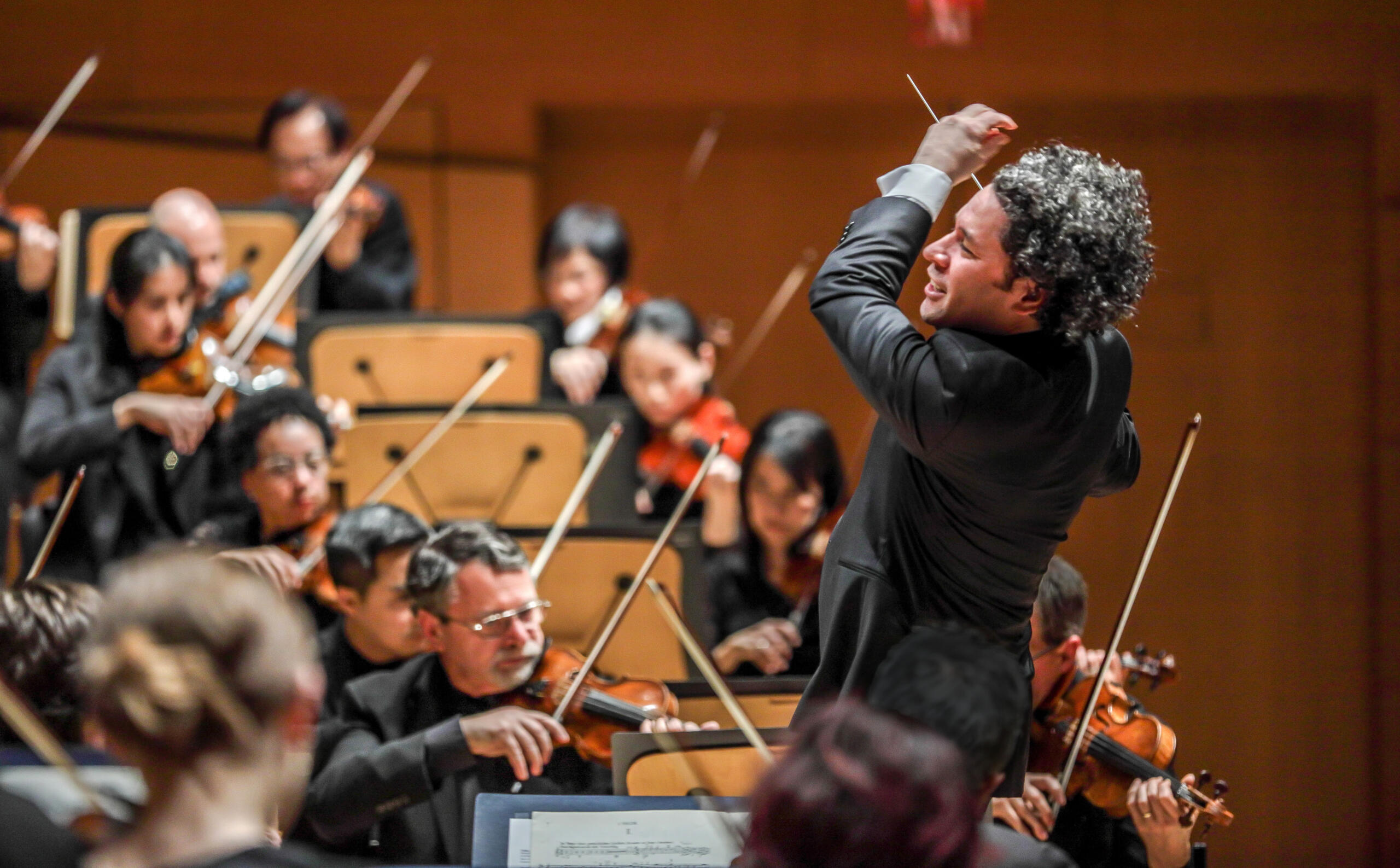 Photograph of the conductor Gustavo Dudamel. He is conducting an orchestra that can be recognised blurred in the background.