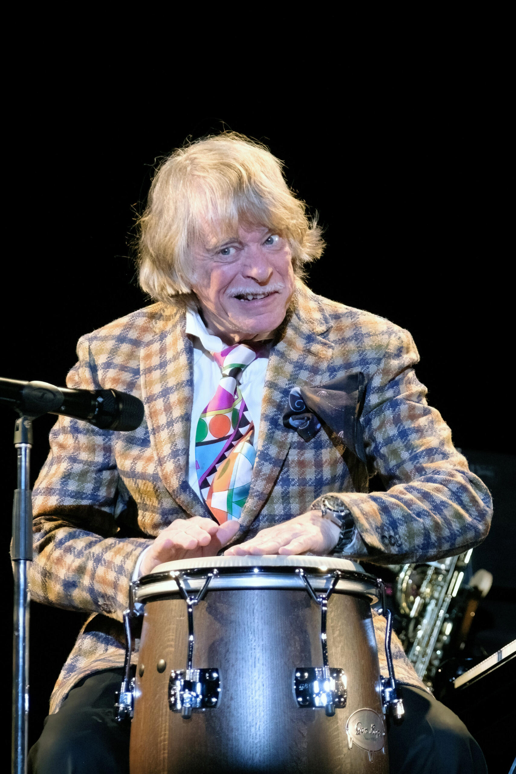 Fotografie des Comedians und Musikers Helge Schneider. Er sitzt vor einem Mikrofon auf einer Bühne und trommelt auf eine Trommel.
