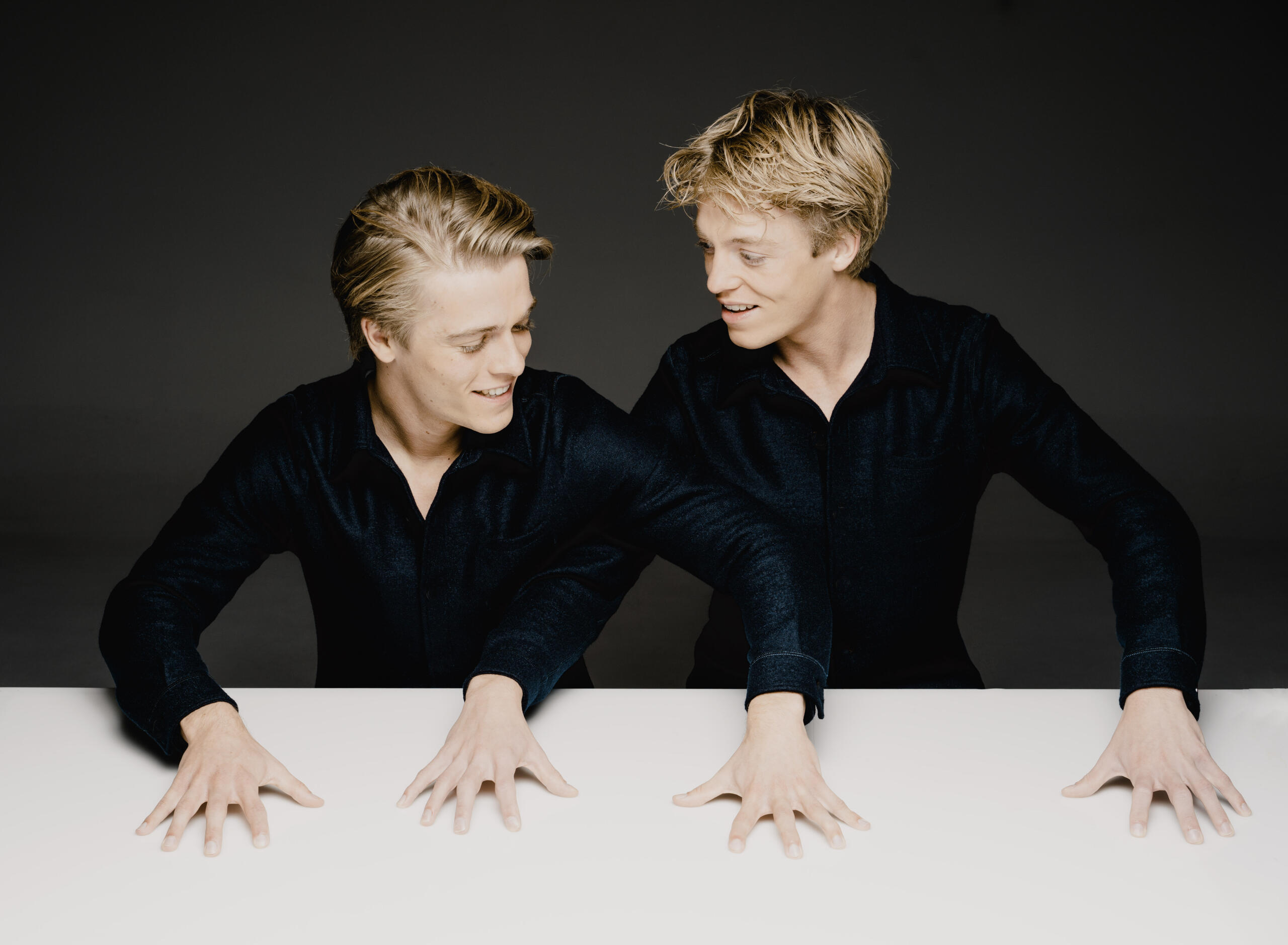 Portrait photograph of the pianists Lucas & Arthur Jussen. The two sit with their hands on the white table in black concert attire in front of a grey wall.