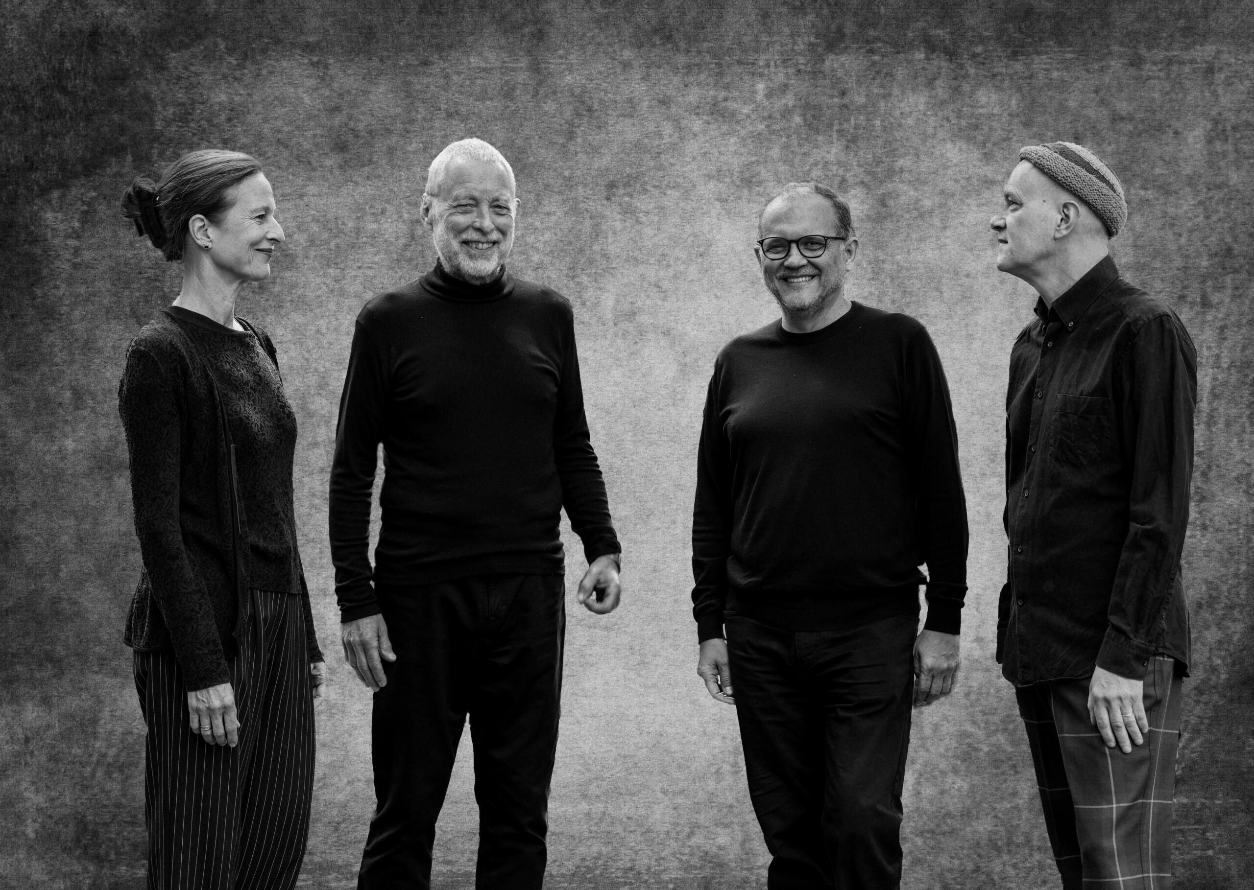 Black and white photograph of three men and a woman dressed in black, standing in front of a grey-mottled wall and smiling at each other.