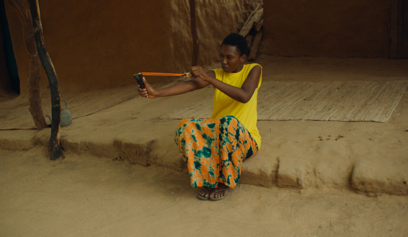 A young person in a yellow shirt and patterned skirt sits on a step and aims a slingshot. White carpets lie on the ground in the background.