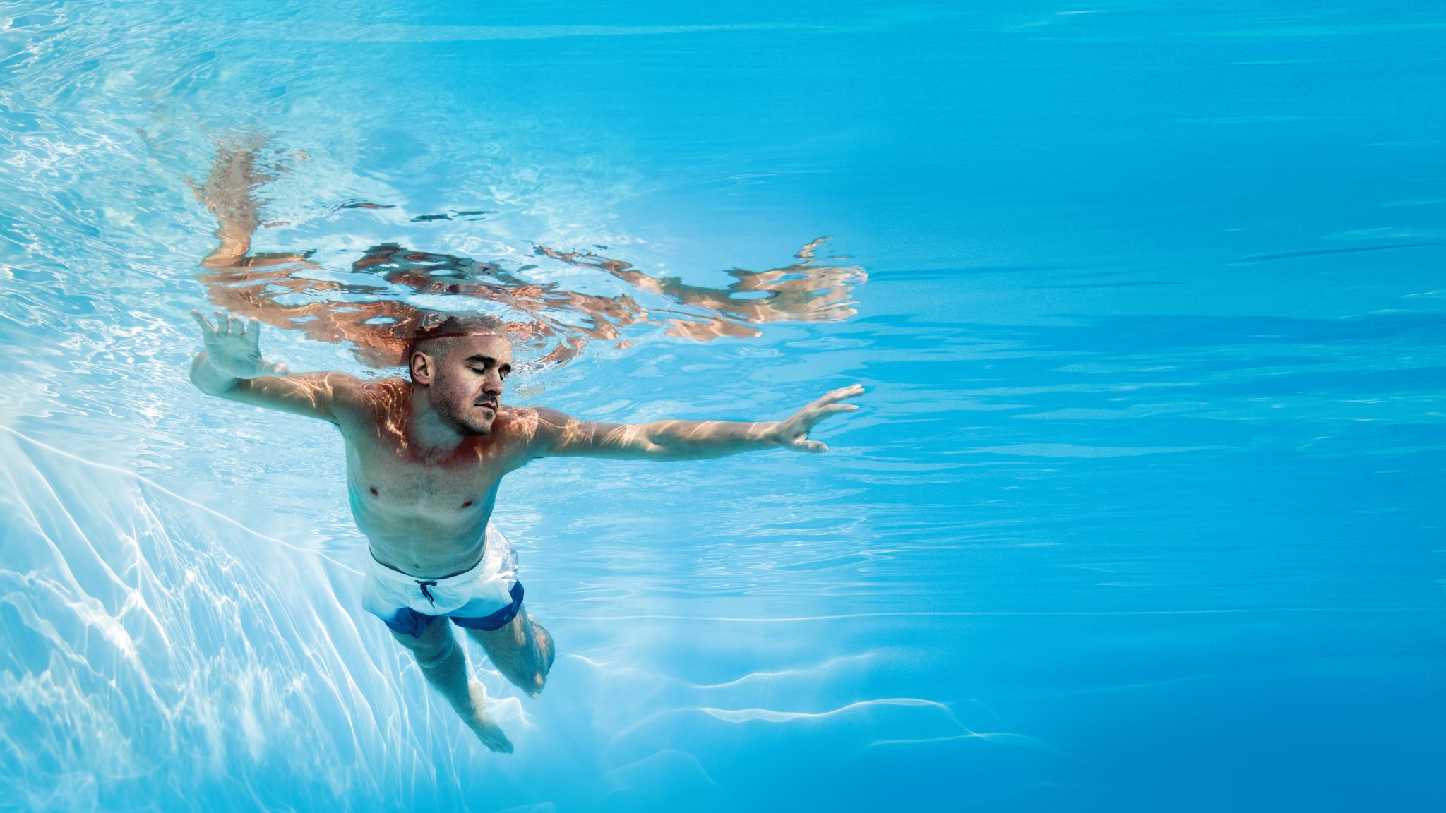 A turquoise-blue swimming pool, you can see a young man swimming from below.