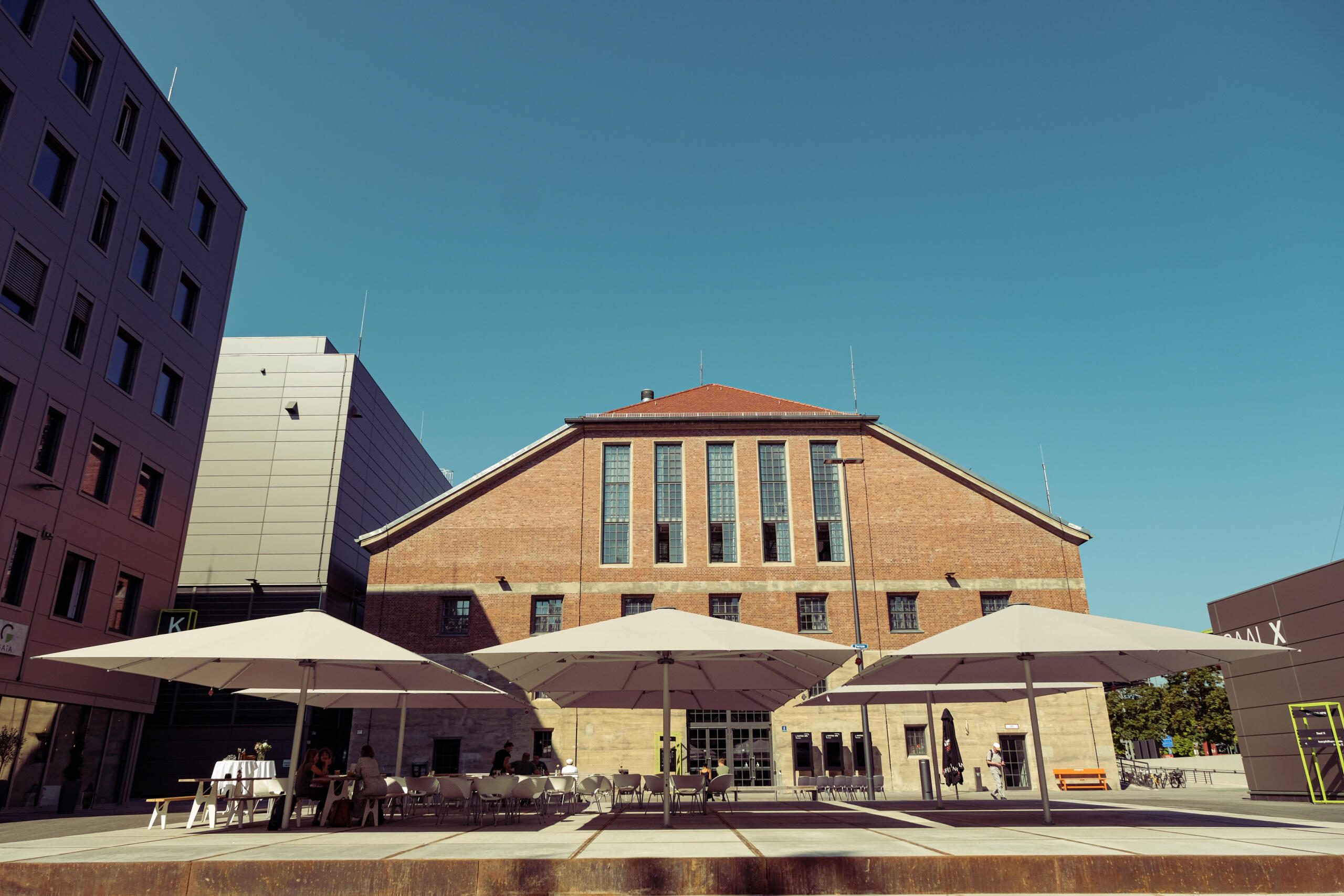 Eine Backsteinhalle, davor sind Tische und Stühle auf einer Freifläche, überspannt von großen Sonnenschirmen.