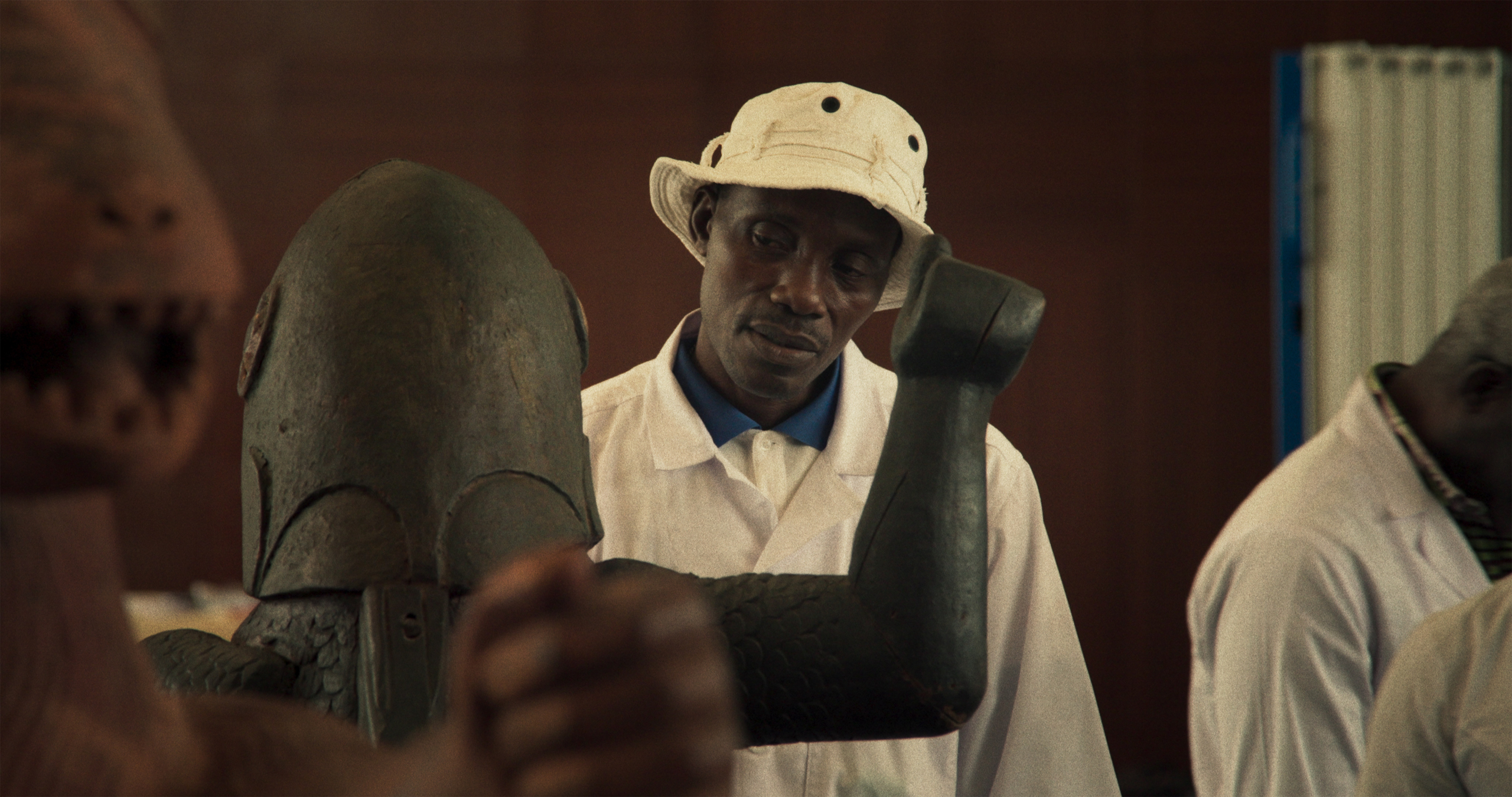 A man in white clothing and a white sailor's hat looks at a statue standing with its back to the camera and raising its right arm.