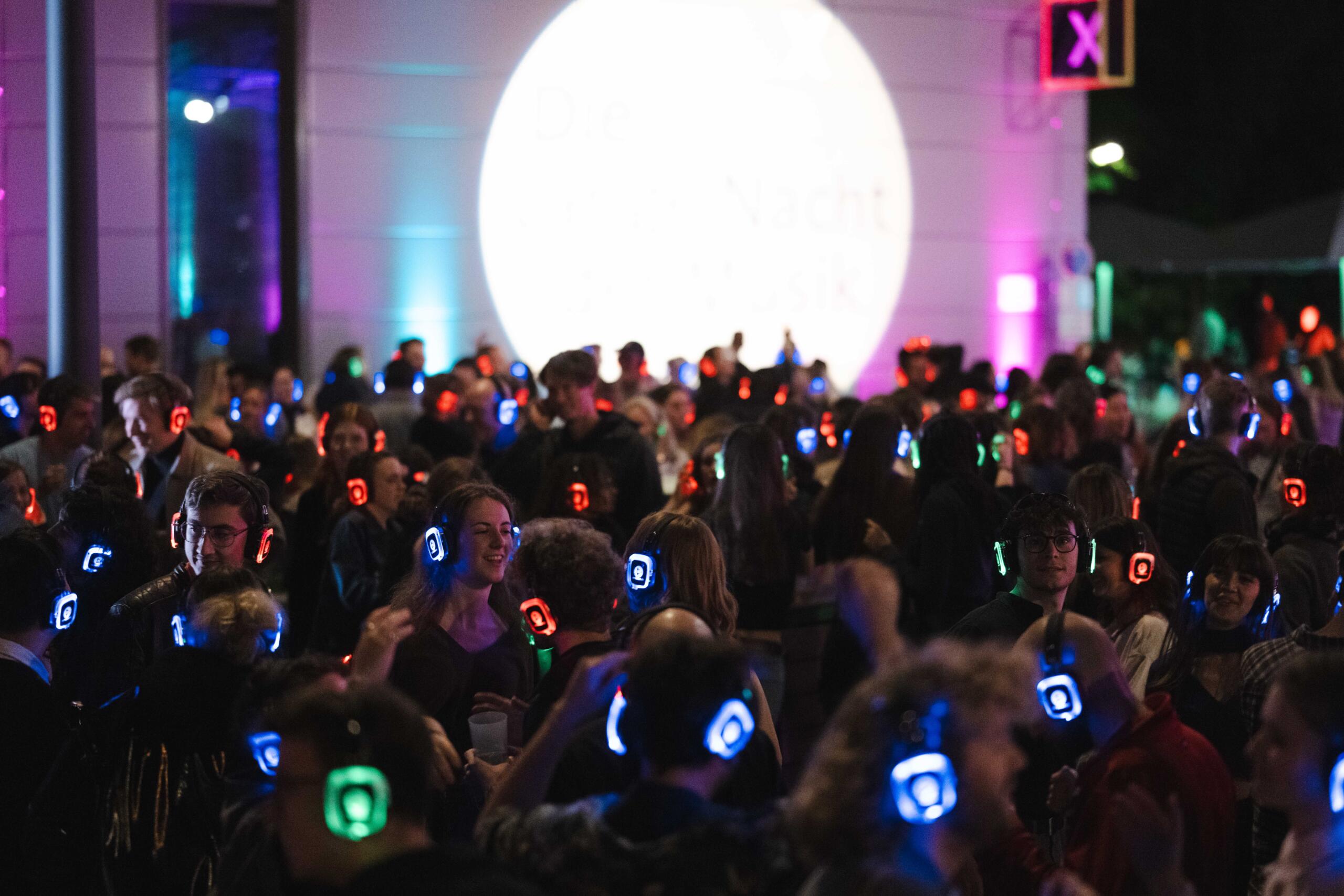 Außenaufnahme, viele Menschen tanzen bei der Langen Nacht der Musik 2024 im Dunkeln mit leuchtenden Kopfhörern auf dem Kopf vor dem Saal X.