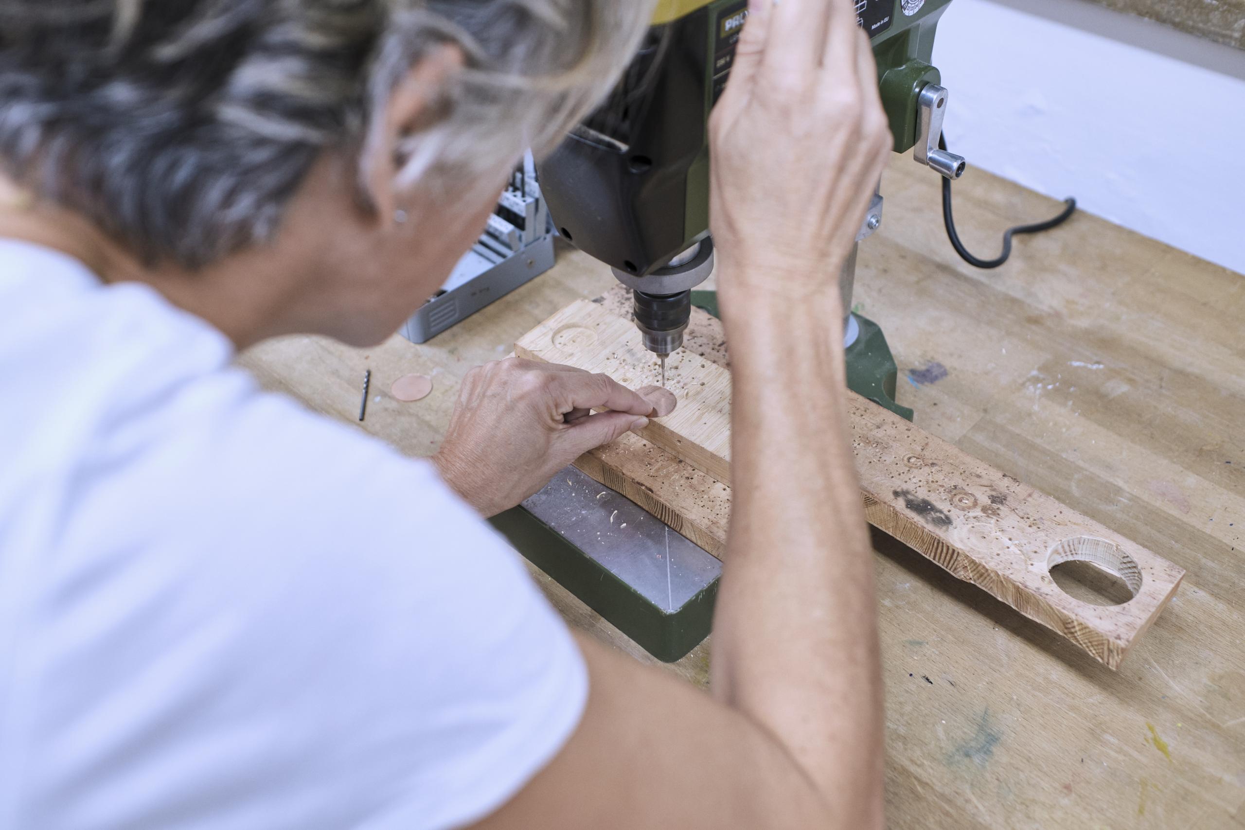 Eine Werkbank, eine Frau nutzt eine Bohrmaschine um ein Metallblättchen zu bearbeiten