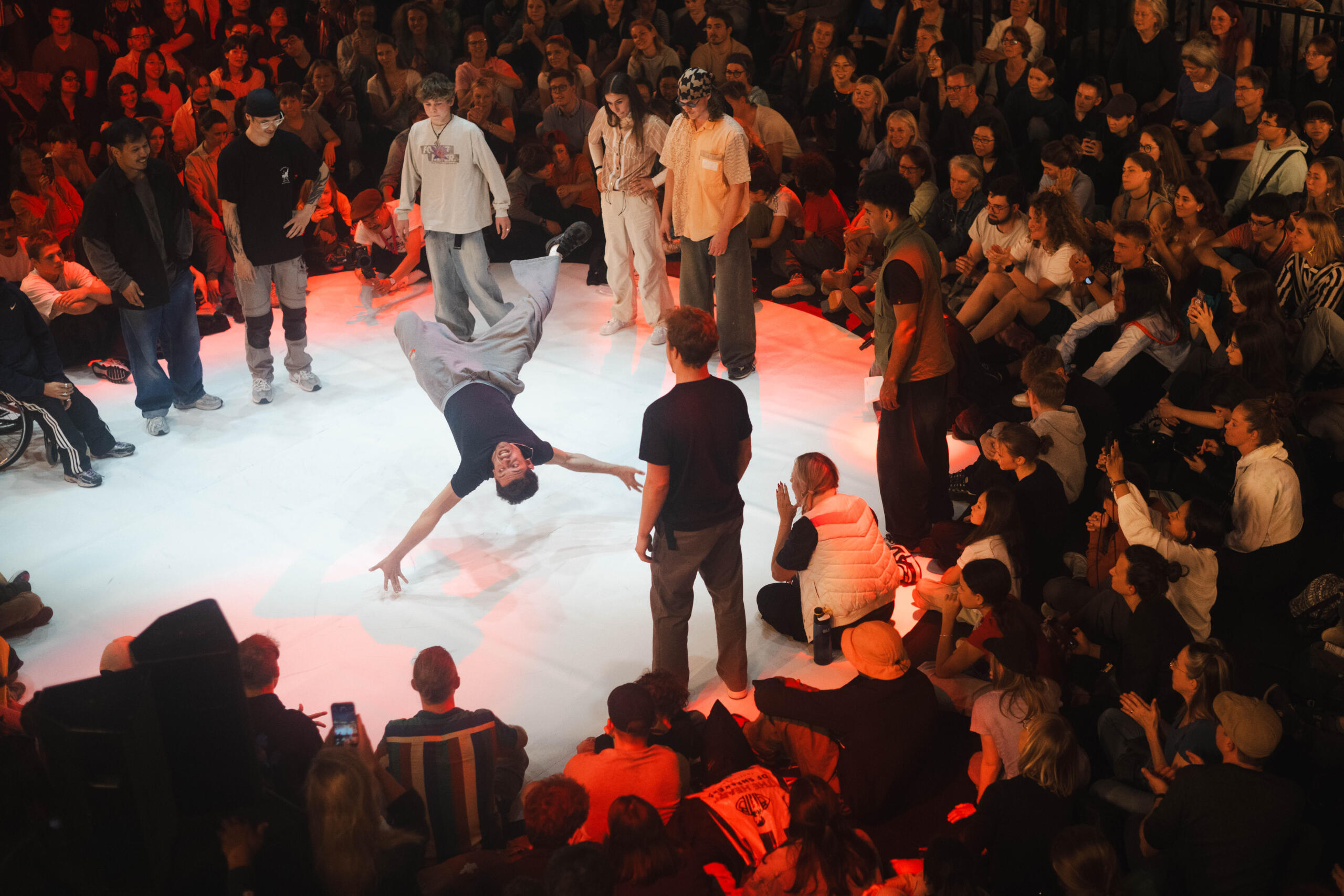 Ein junger Mann fliegt beim Breakdance durch die Luft, um ihn herum sitzen und stehen Zuschauende im Kreis.