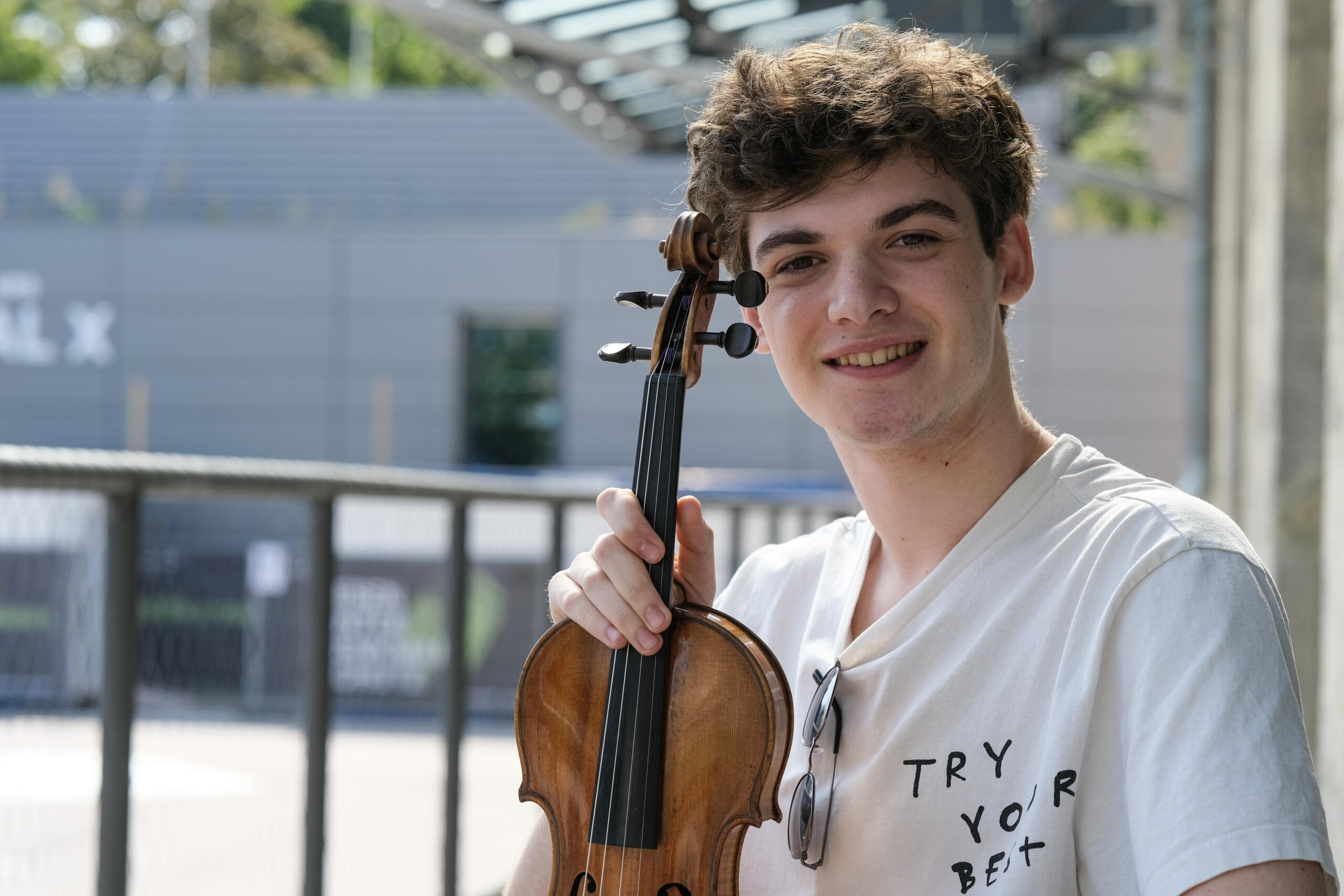 A young man on an outdoor terrace, laughing into the camera and holding up a violin. His T-shirt reads "Try your best".