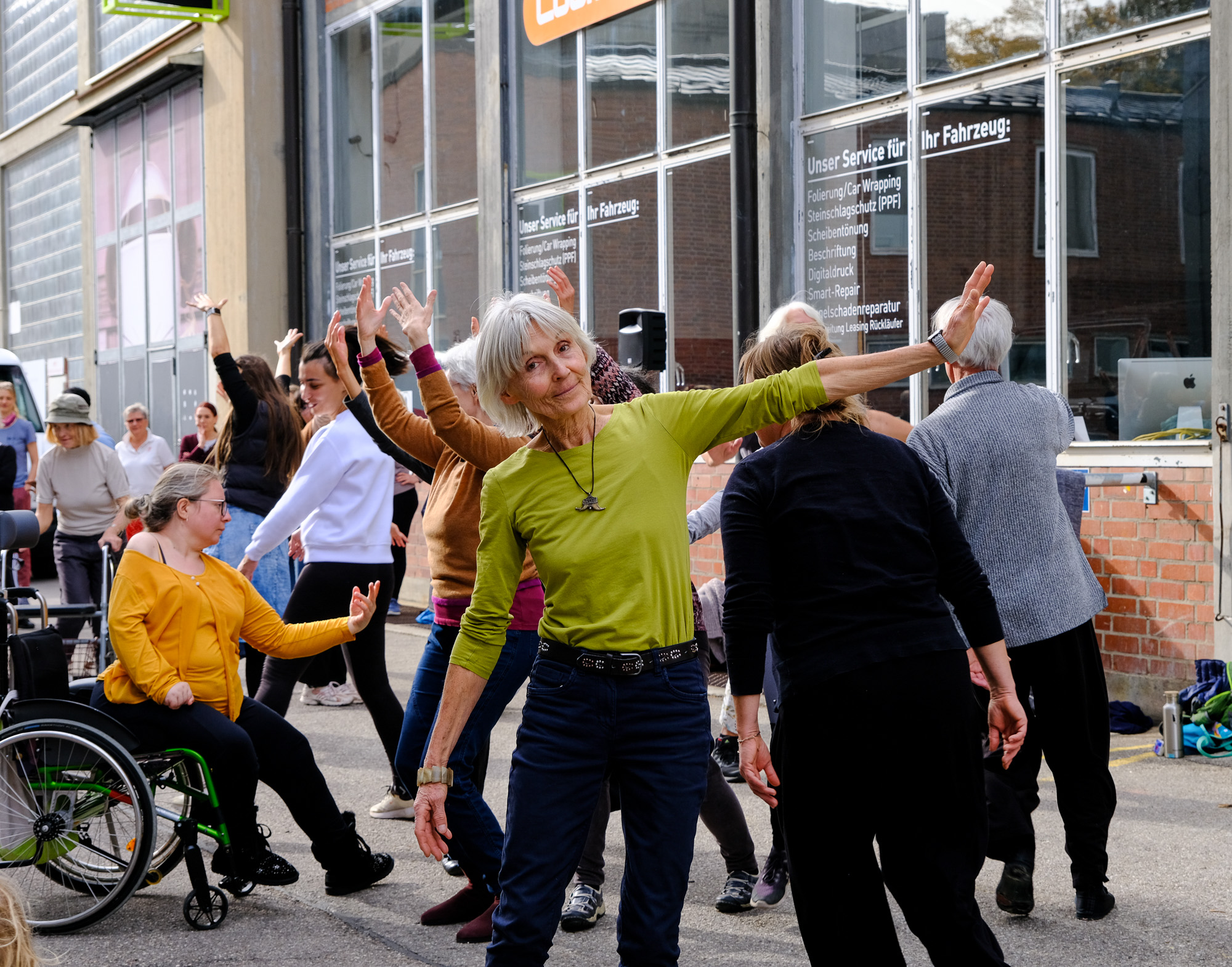 Outdoor area, daytime, a group of people of all ages dance with individual movements on the grounds of Gasteig HP8, one lady dances along sitting in a wheelchair.