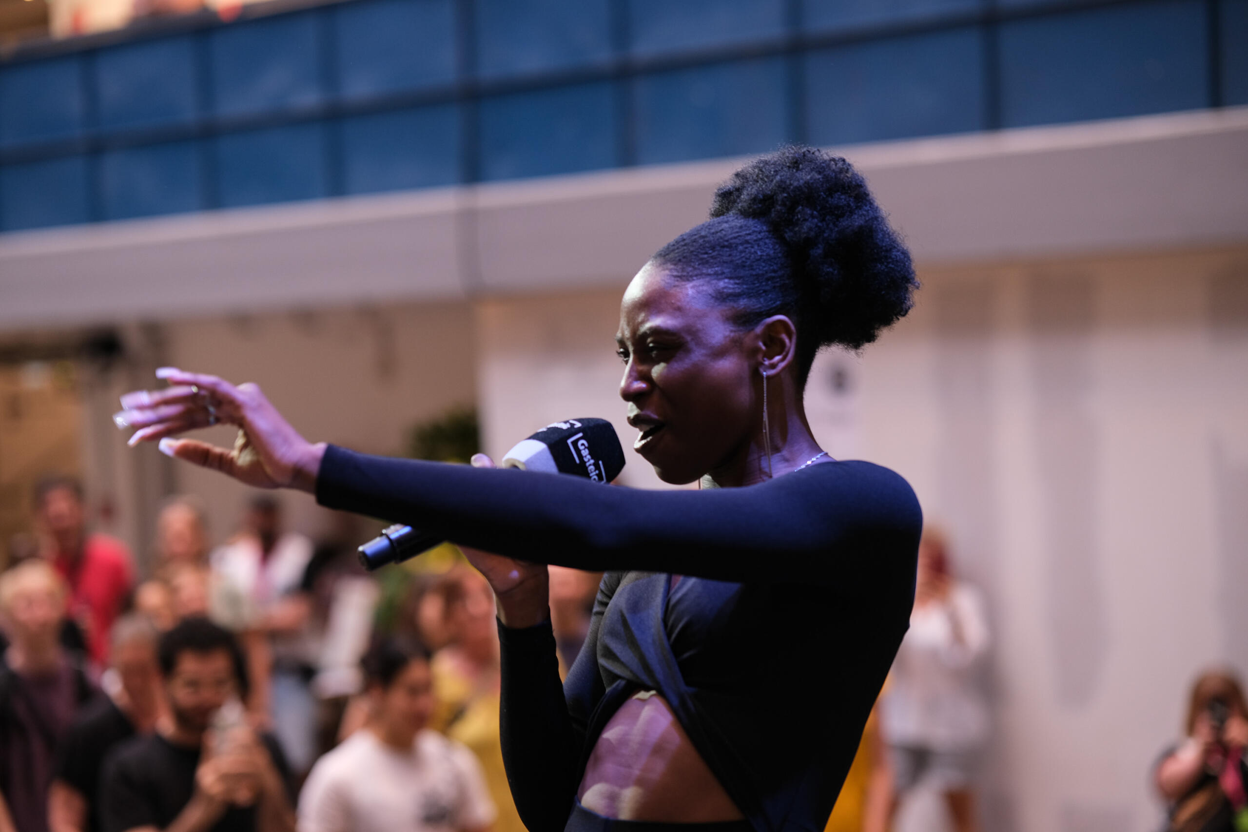 A black woman sings into a microphone and stretches one arm forward.