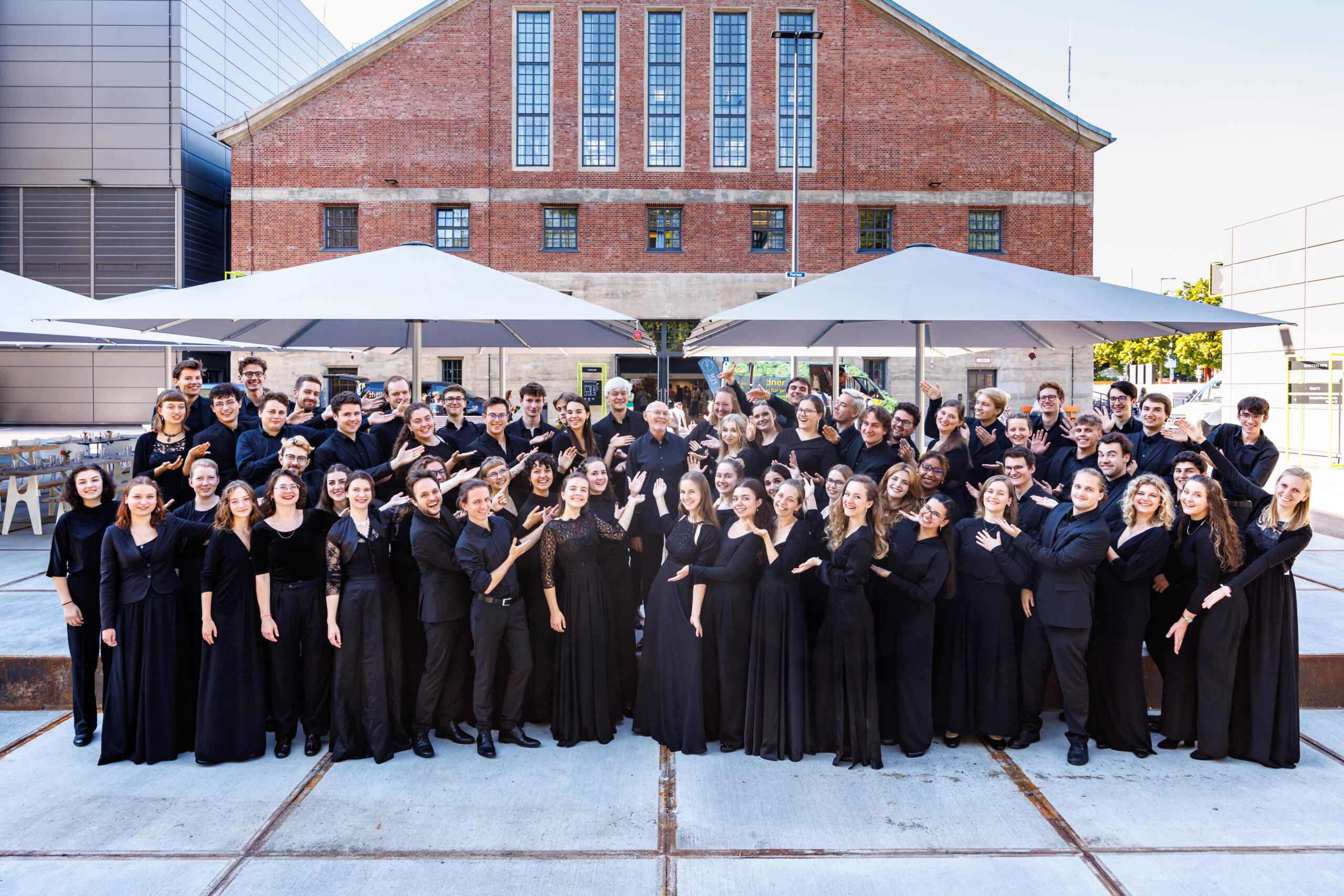 A large choir of young people dressed in black stands on a terrace, with the brick façade of Hall E in the background.