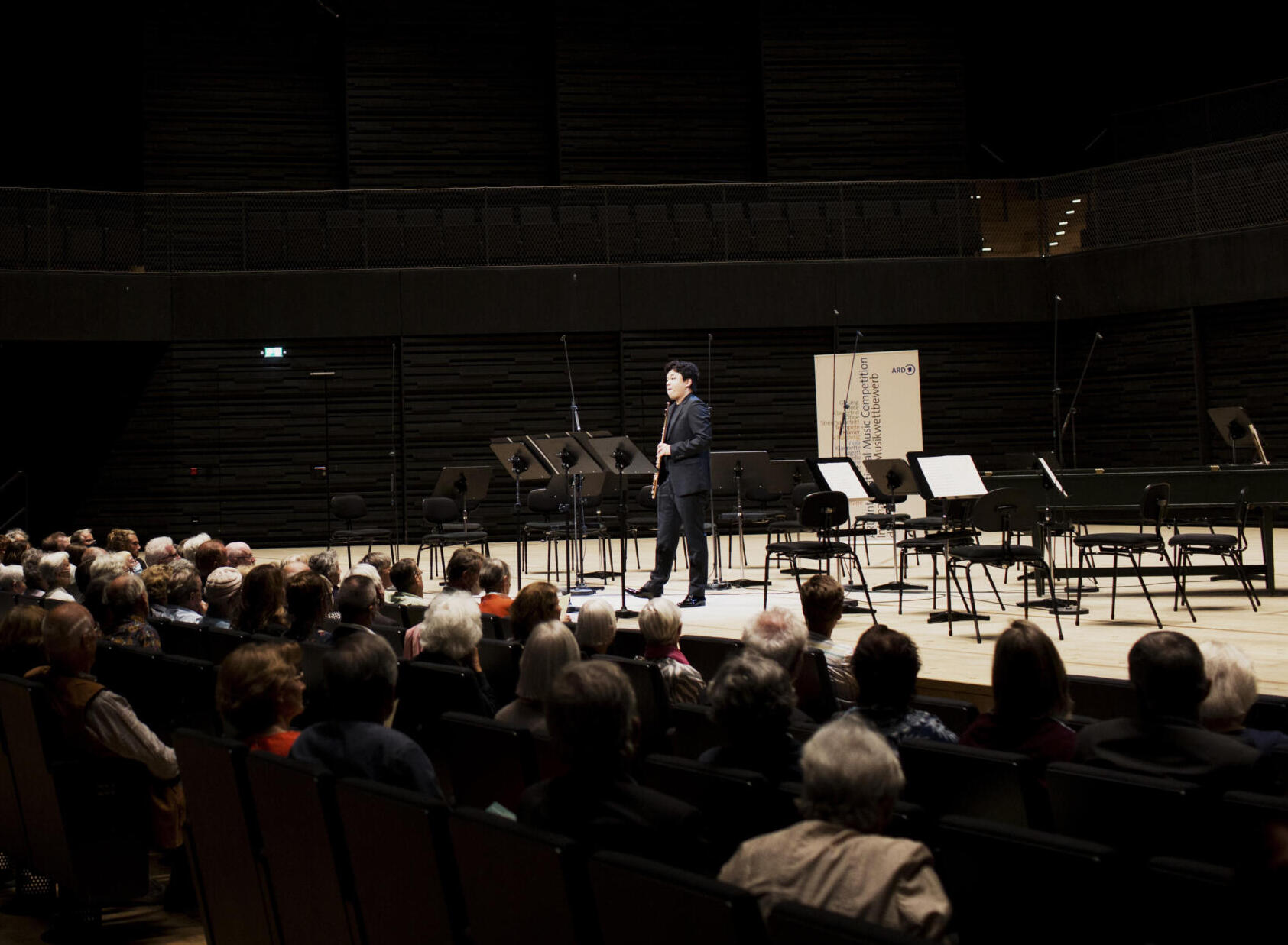 A musician stands at the front of the brightly lit stage of the Isarphilharmonie. The silhouettes of the audience can be recognised in the dark.