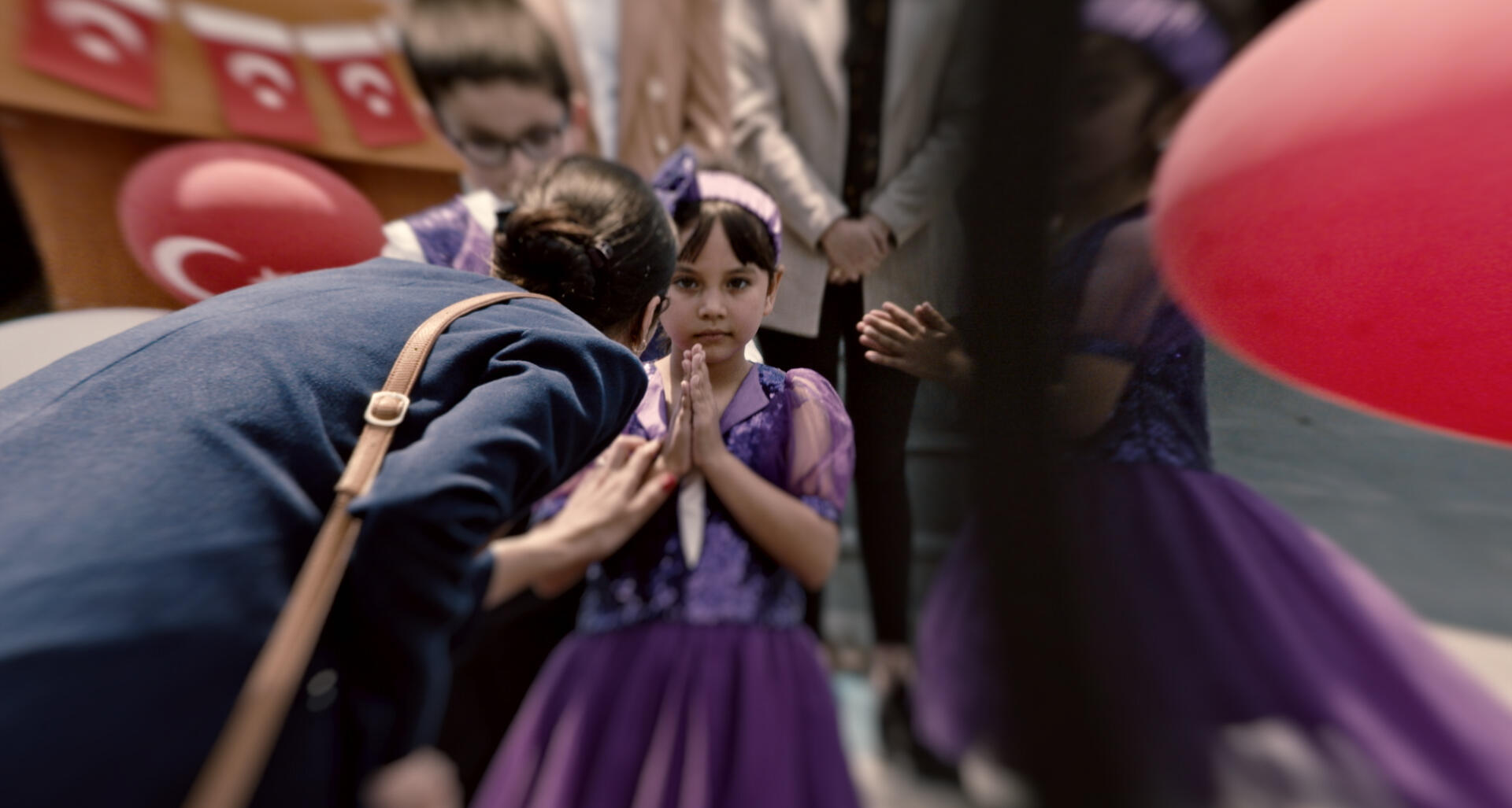 A little girl in a purple dress stands next to a woman and puts her palms together. The perspective of the picture is distorted.