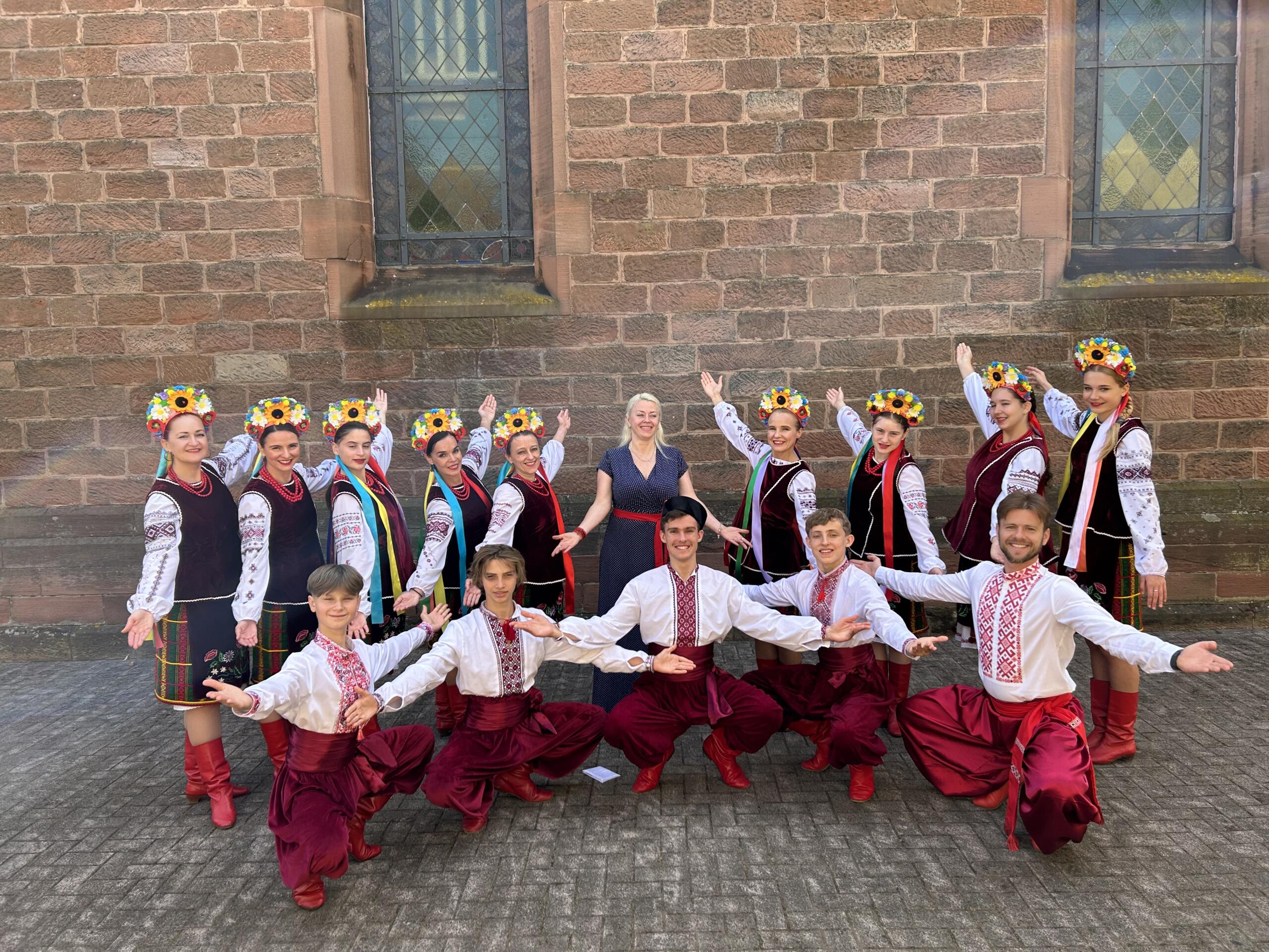 A group of people in front of a brick wall, wearing folkloric clothing, the women wearing wreaths of flowers in their hair.