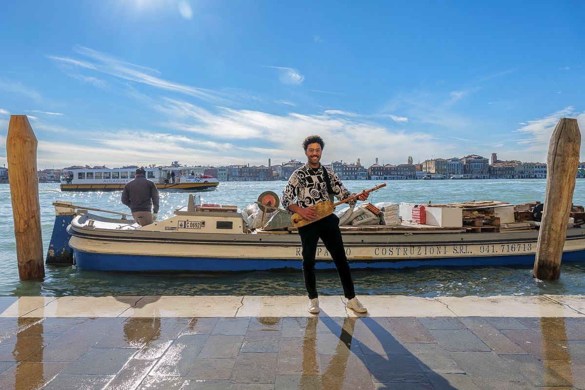 A man with a stringed instrument stands by a canal, behind him is a cargo boat, another boat and houses on the other side of the canal in the distance.