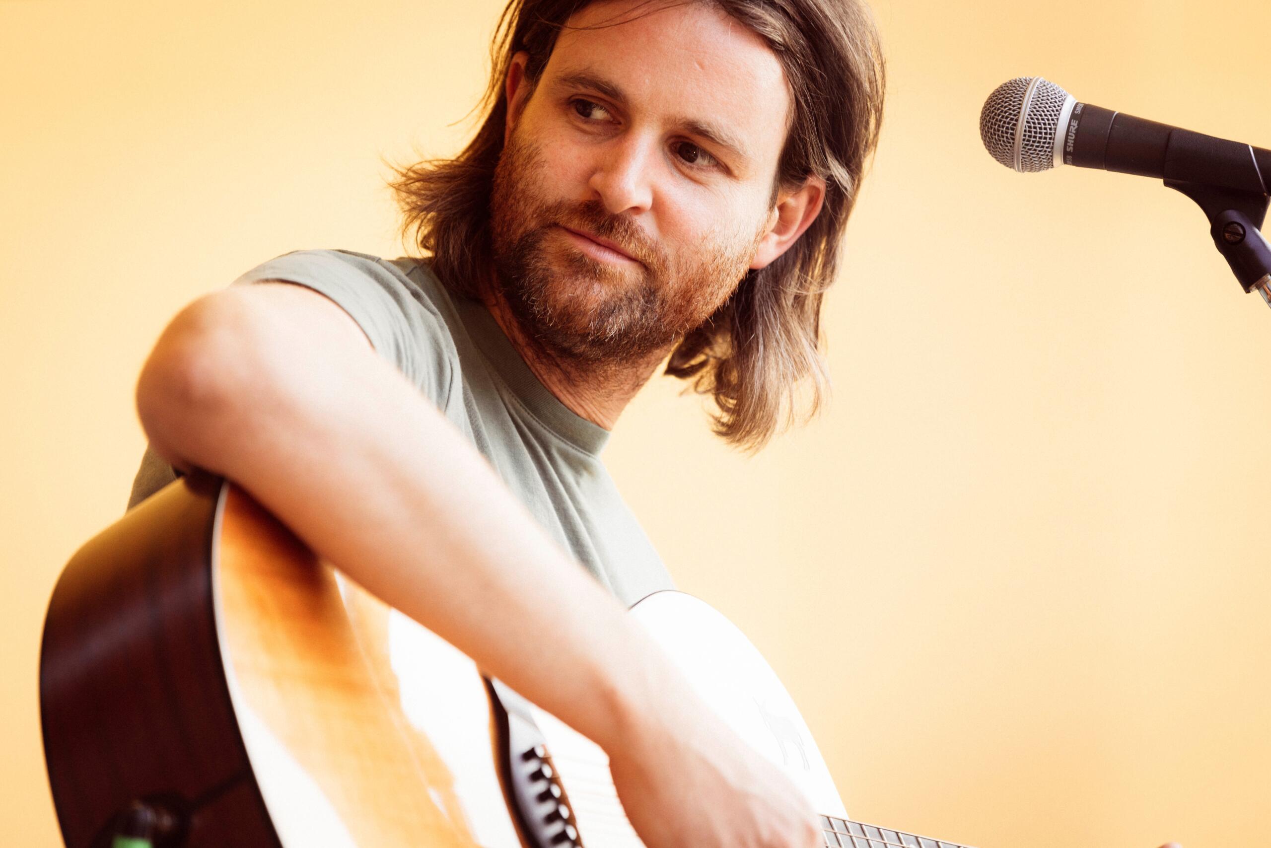 A man with half-length hair and a beard plays the guitar.