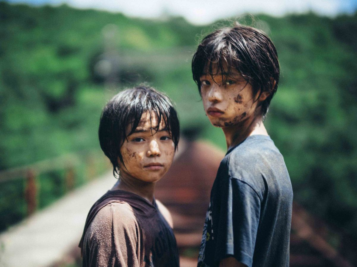 Two young boys are standing on a bridge, which can be seen blurred in the background. They both have mud on their faces.