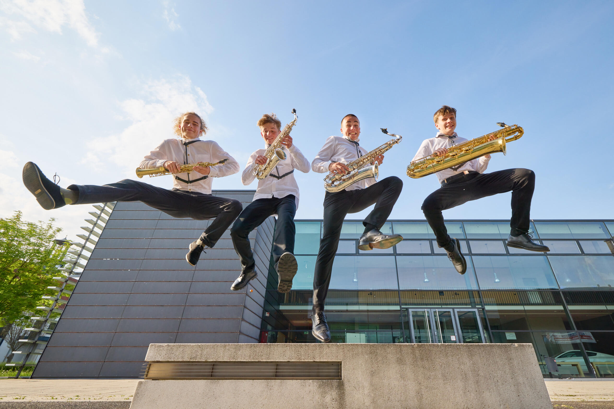 Vier jugendliche Musiker springen von einer Rampe. Sie tragen Konzertkleidung und halten ihre Saxophone in den Händen.
