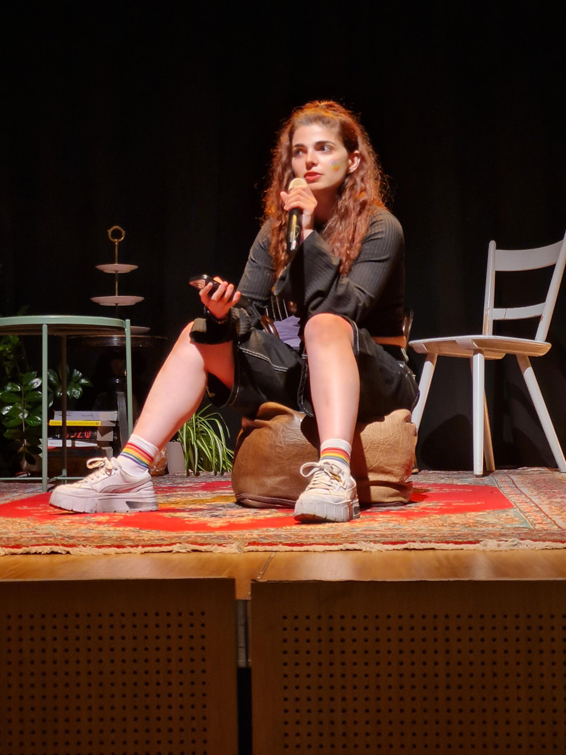 A young woman with long curls is sitting on a stool, surrounded by various pieces of furniture and decorations. She is holding a mobile phone and speaking into a microphone.