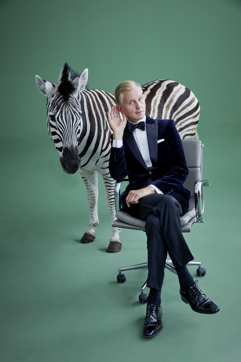 Max Raabe sitzt in Anzug vor einem grünen Hintergrund auf einem Schreibtischstuhl und hinter ihm steht ein Zebra.