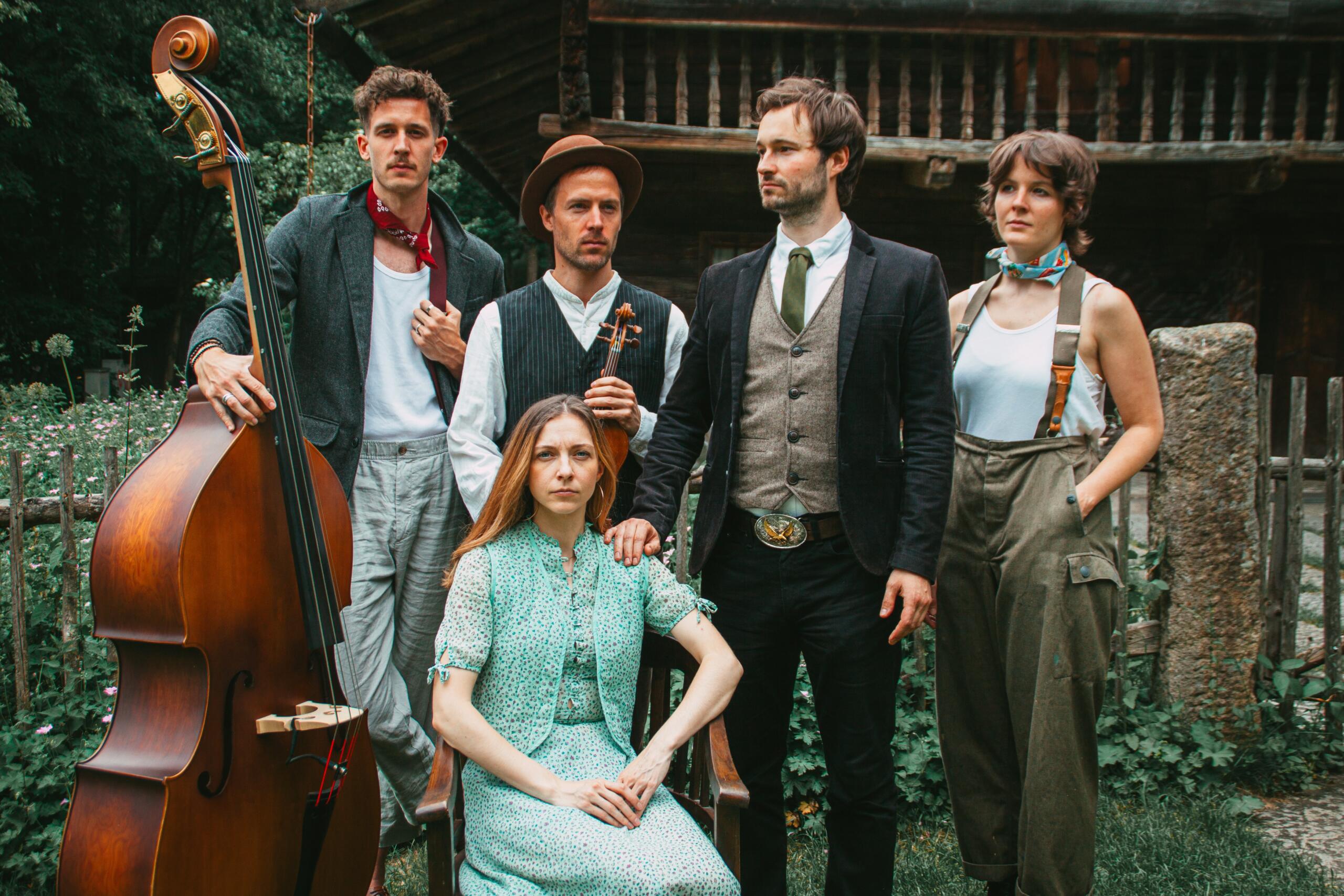 The five members of the band are standing in front of a historic farmhouse.