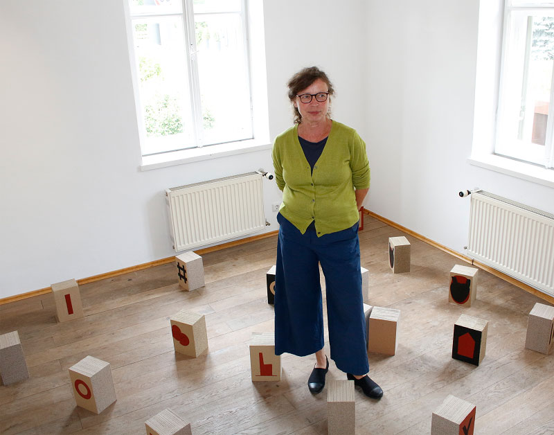 The artist Susanne Pittroff stands in the corner of a room with small wooden blocks with symbols scattered around her.