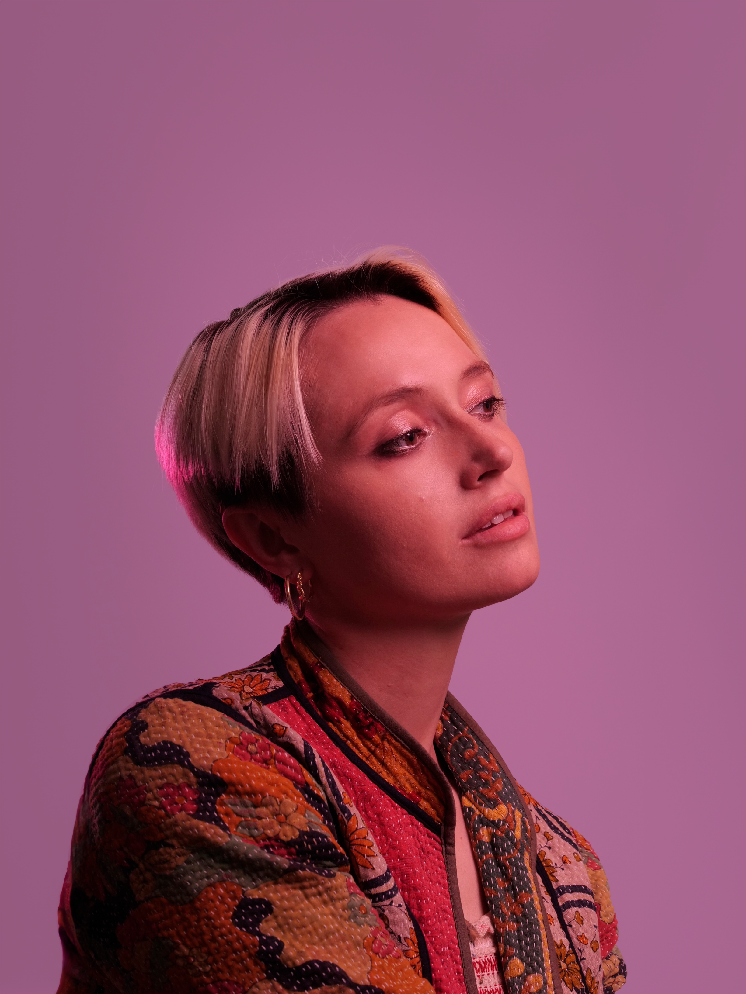 Portrait of the musician Sarah Reema Müller-Westernhagen. A young woman with short blonde hair looks thoughtfully to the right. The background is pink.