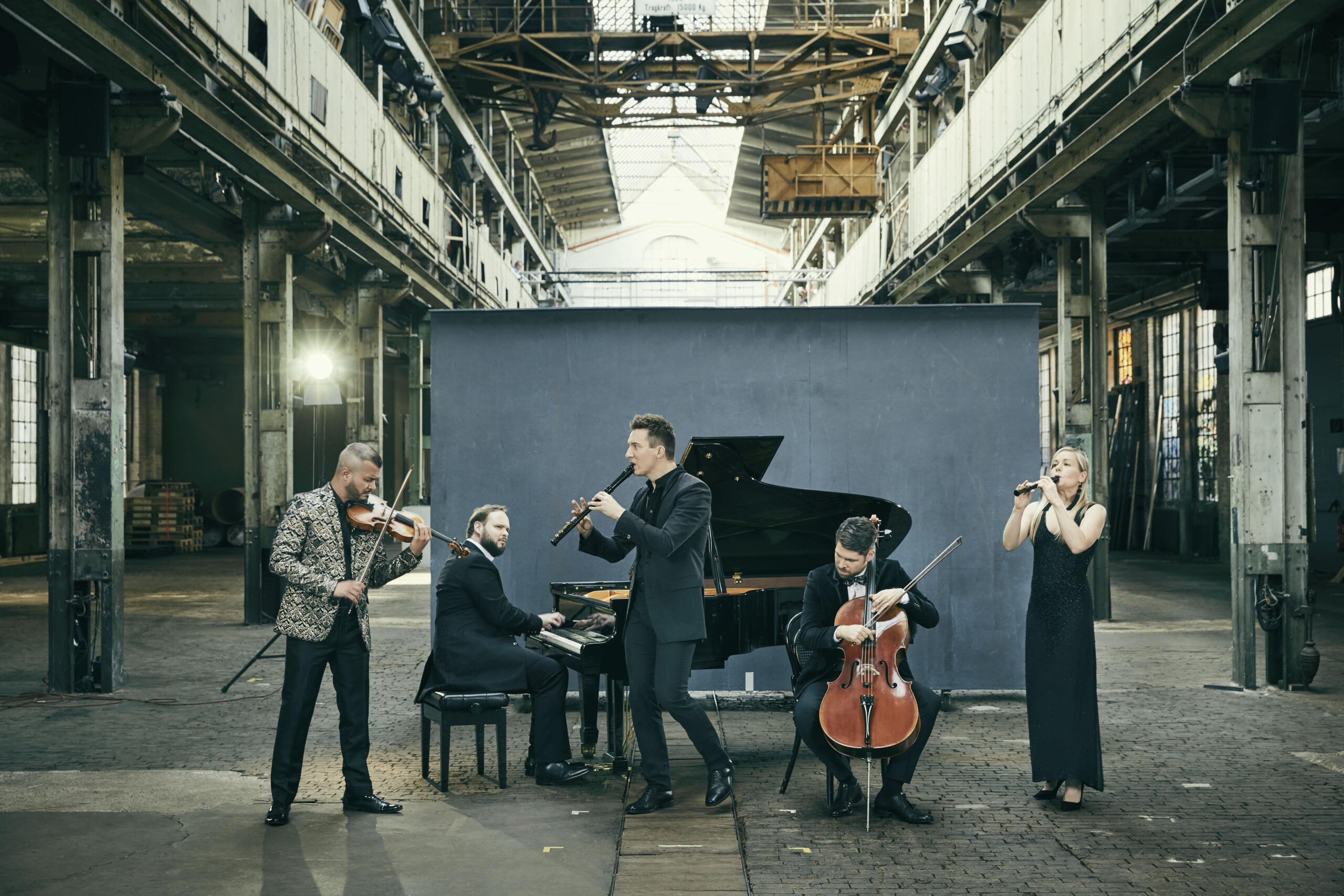 Four musicians and a female musician stand in an abandoned machine hall and play music. A grey screen is set up behind them.