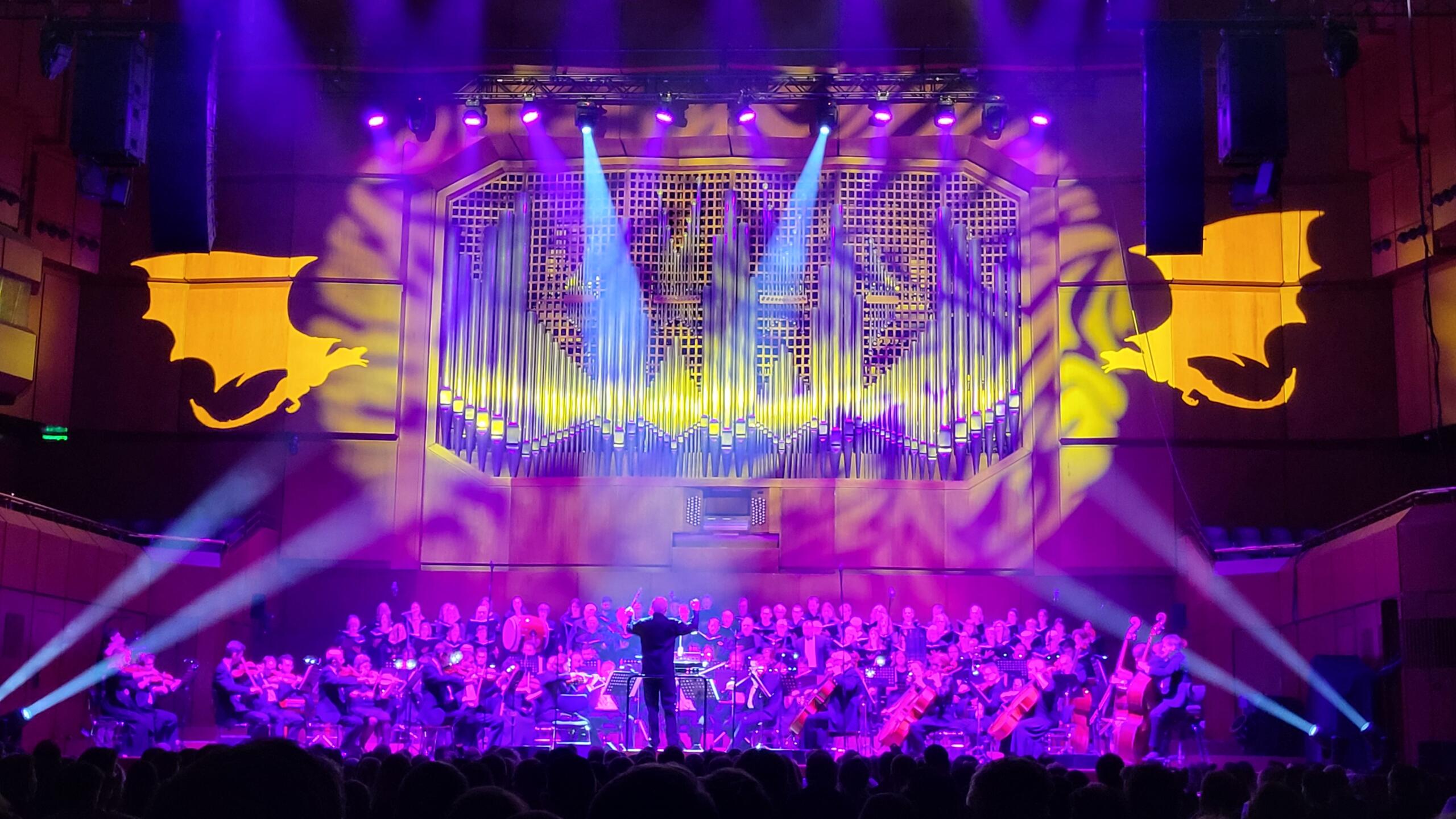 An orchestra sits on a colourfully lit stage. Two dragons spitting fire are projected onto the wall behind it.