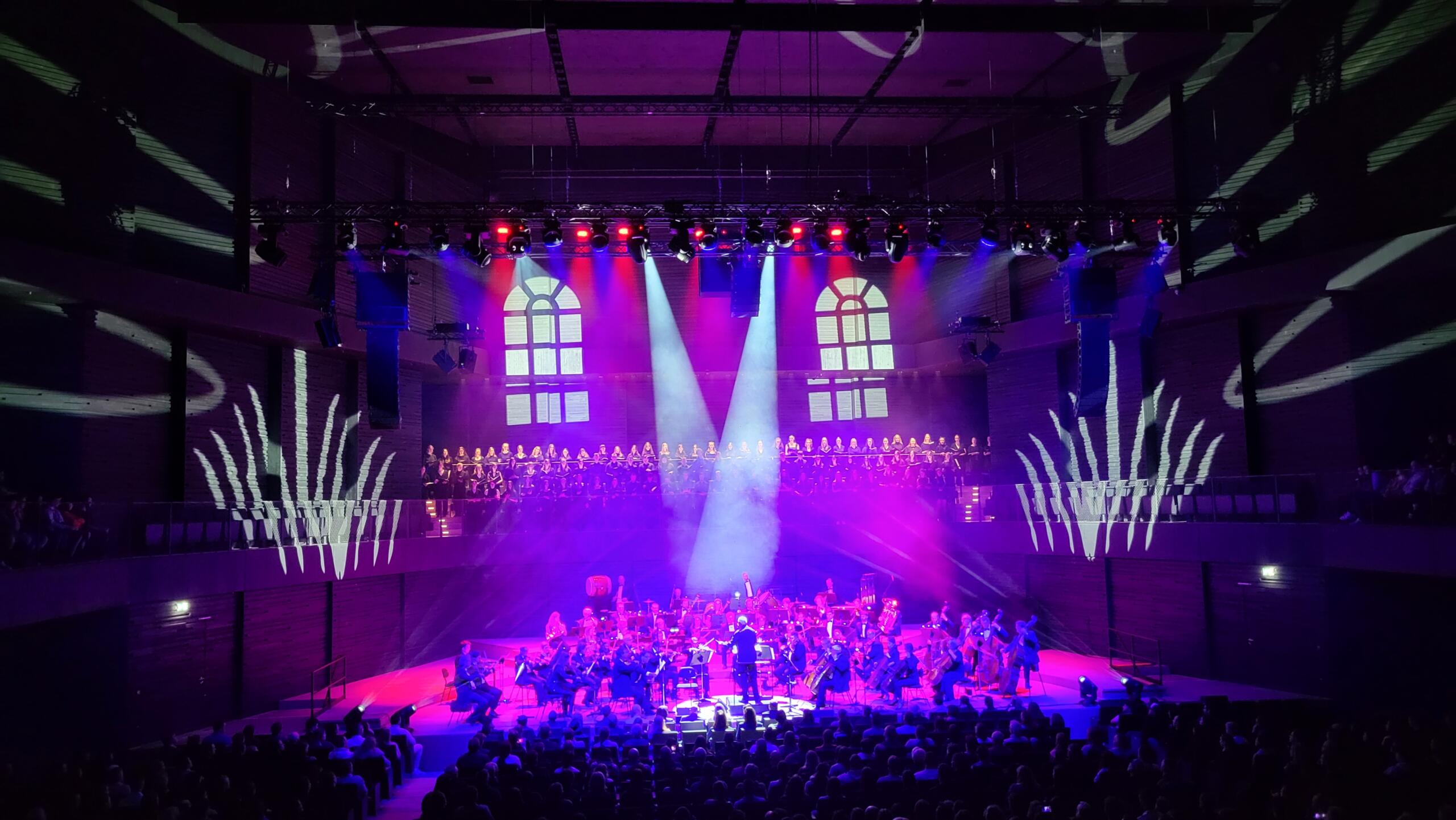 Ein Orchester spielt auf einer bunt erleuchteten Bühne. Dahinter steht ein Chor auf einer Empore. An beiden Seiten sind weiße Projektionen zu sehen.