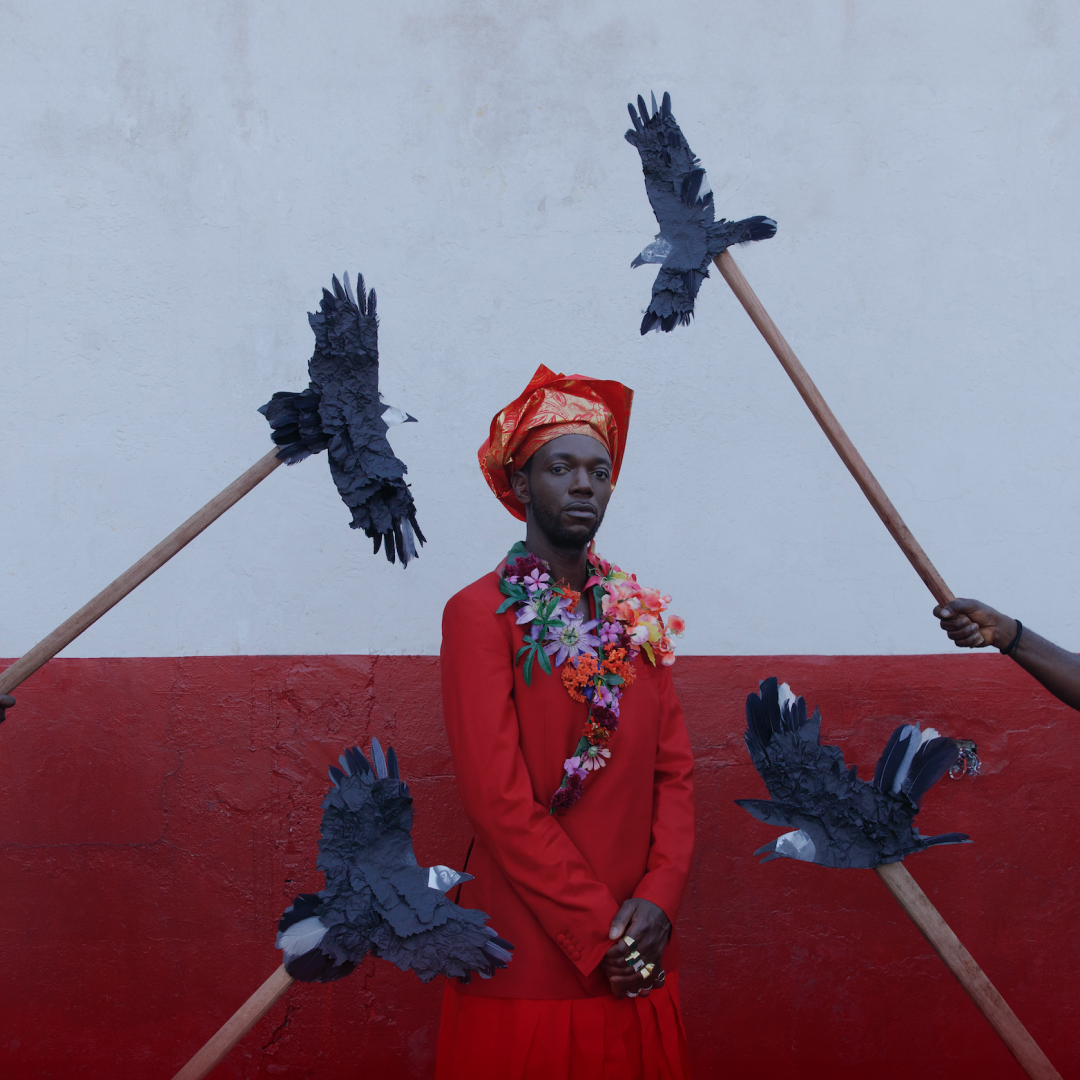 A black man in a red robe stands in front of a red and white wall. Black paper birds on sticks are held up in the air around him.