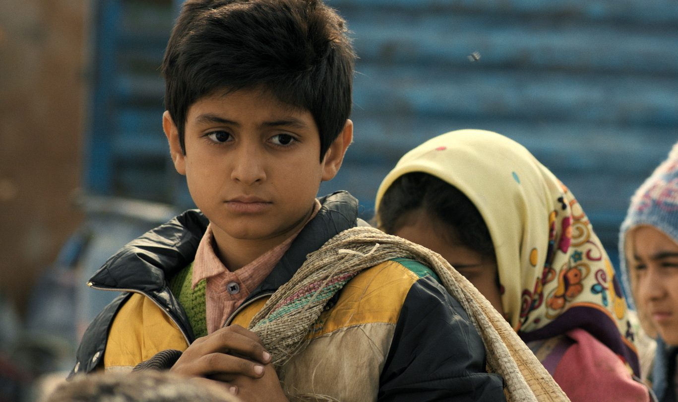 A small boy with black hair and dirty clothes looks seriously and sadly into the distance.