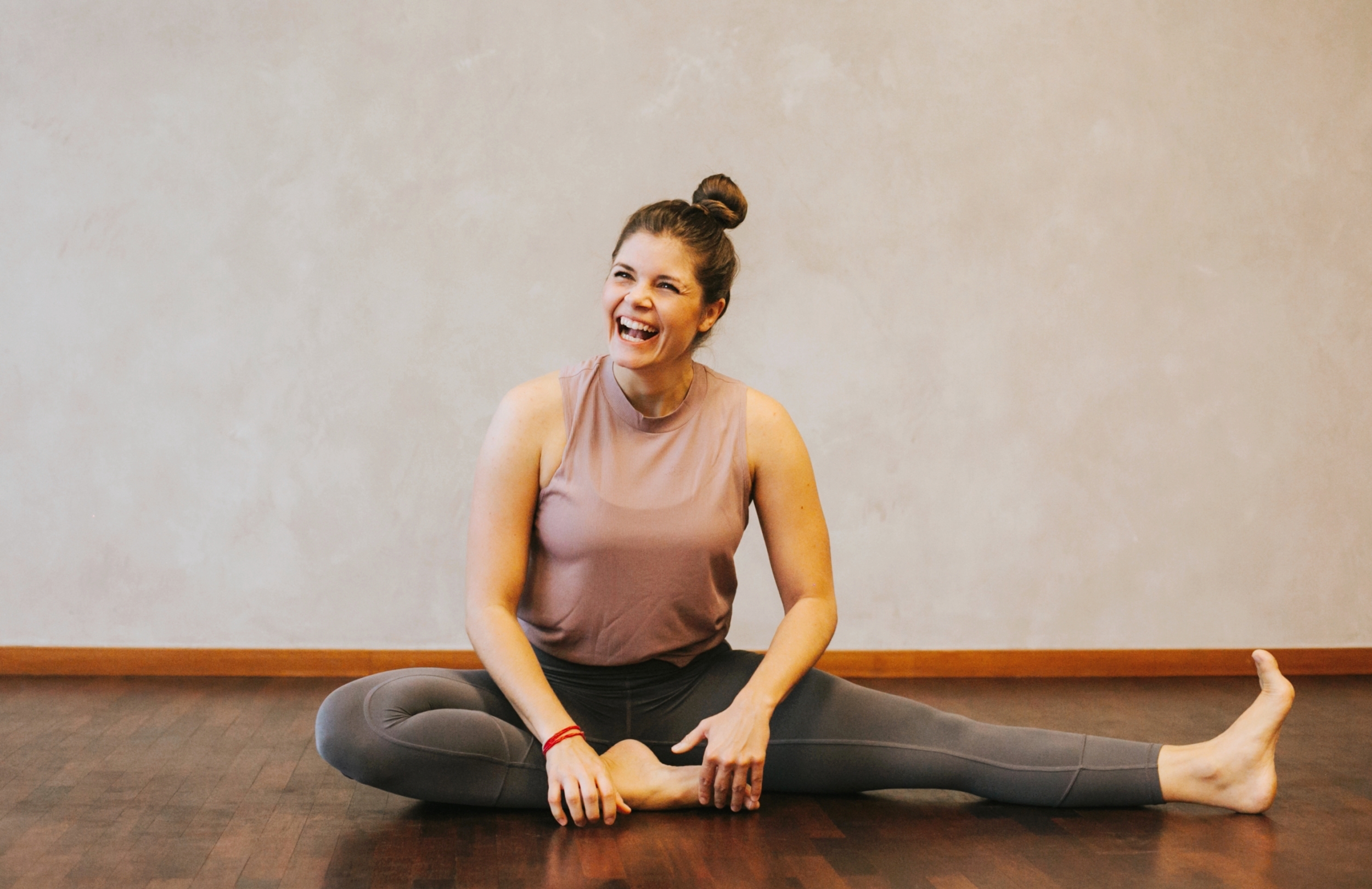 A woman sits laughing in a yoga pose on a parquet floor.