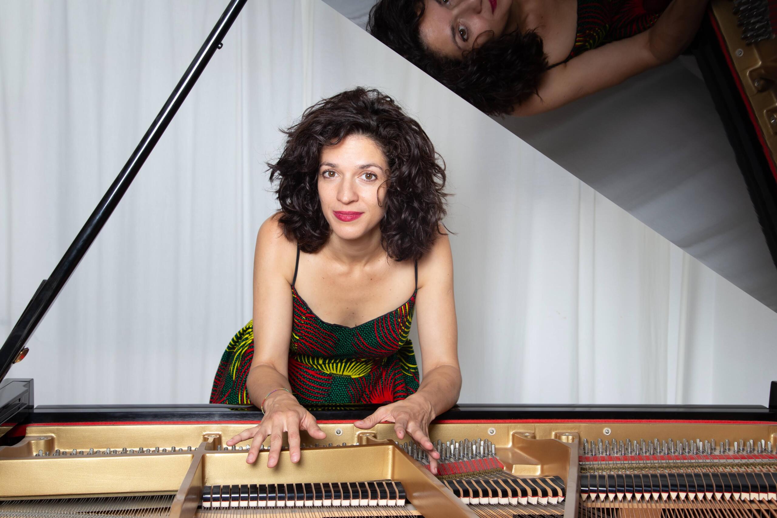 Portrait photograph of Cuban pianist Yami CruzMontero. She stands behind a grand piano in a colourful dress and smiles at the camera.