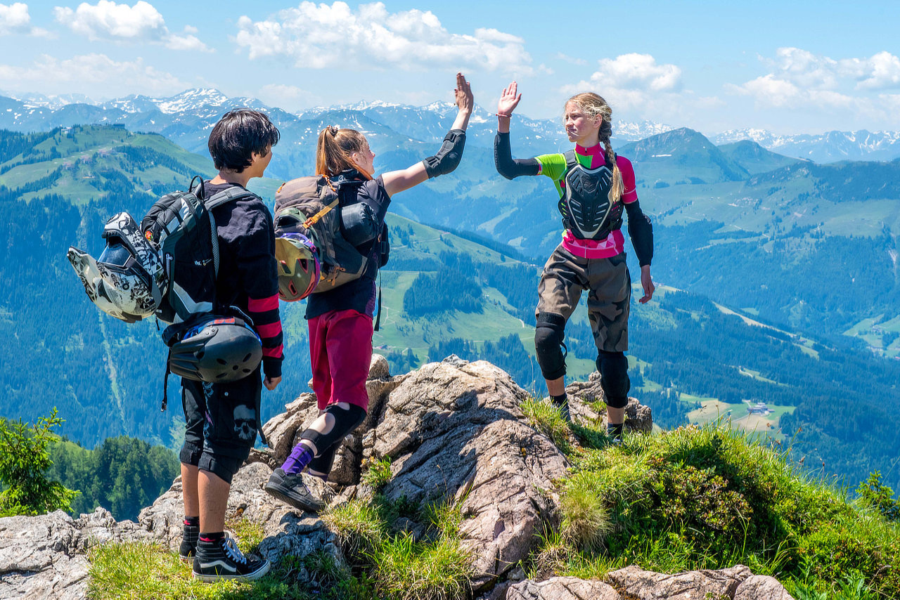 Drei Jugendliche stehen in Mountainbike-Kleidung auf dem Gipfel eines Berges und klatschen sich ab. Im Hintergrund sieht man eine Bergsilhouette.