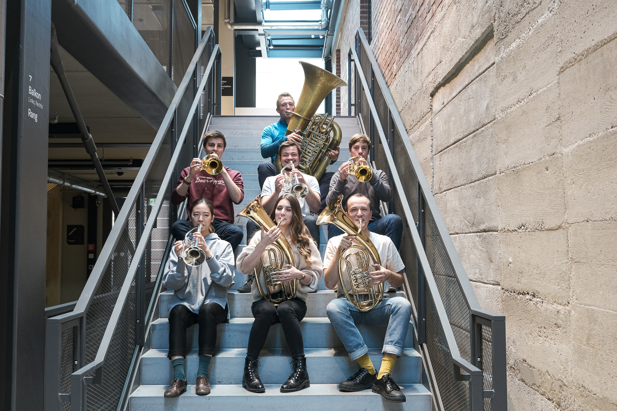 7 Studierende mit ihren Instrumenten sitzen auf den Stufen des Treppenhauses der Isarphilharmonie und machen Musik